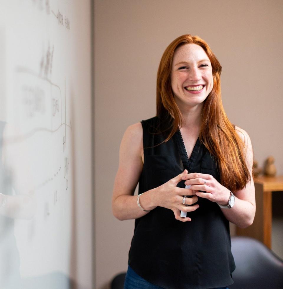 Smiling woman with long hair wearing a black dress and standing in front of a whiteboard