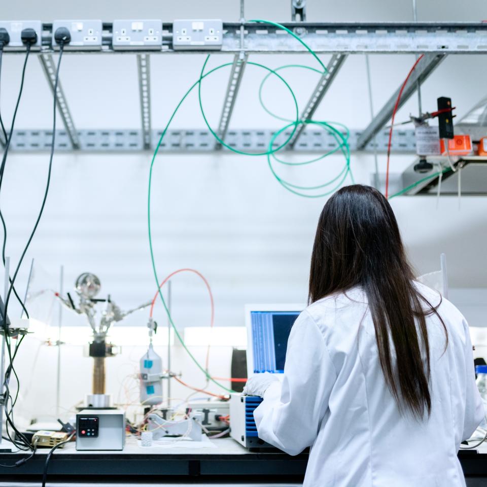 Female engineer in white coat working at a lab bench, with back to camera