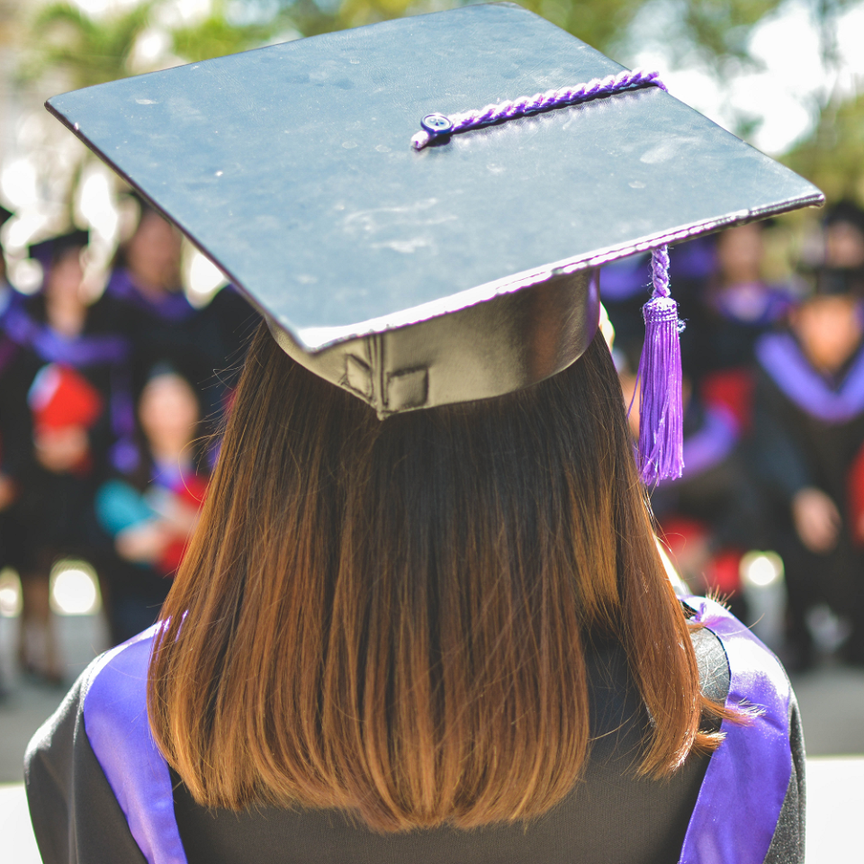 A student in graduation gowns standing in front of a seated crowd of students in graduation gowns.