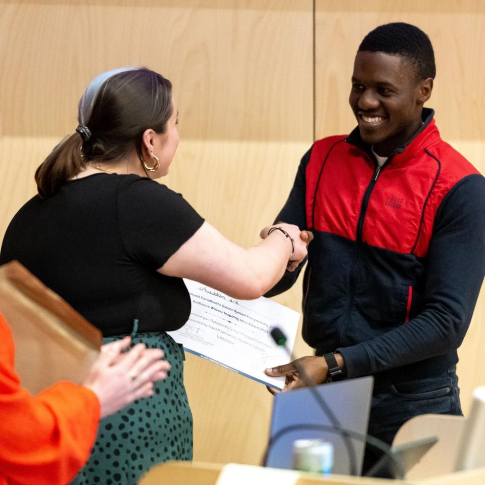 A woman shakes hands with a young man while handing him a certificate. Another woman applauds in the background