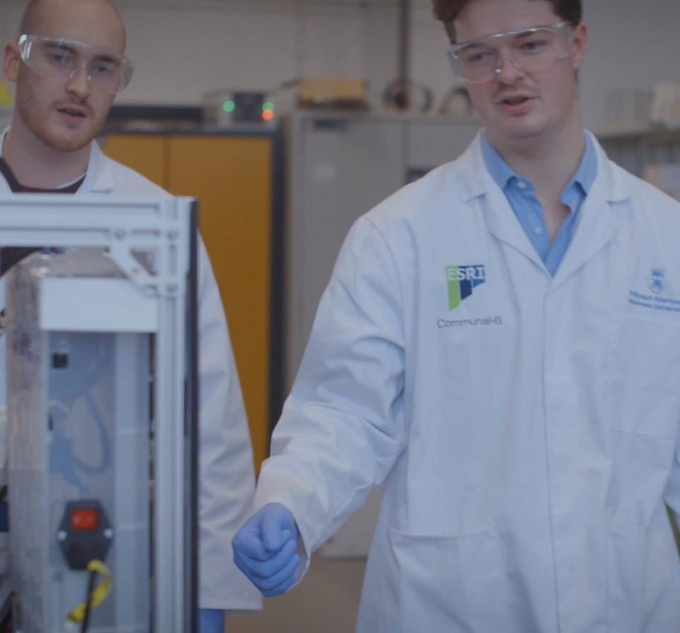 Two men wearing white lab coats, blue gloves and protective goggles, standing in front of a piece of equipment