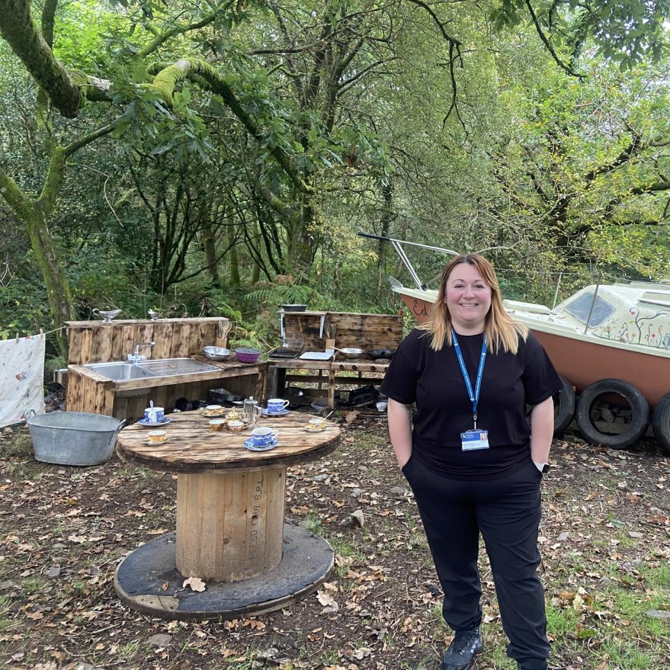 A woman dressed in black standing in a forest setting with wooden benches, wooden table, and a boat behind her