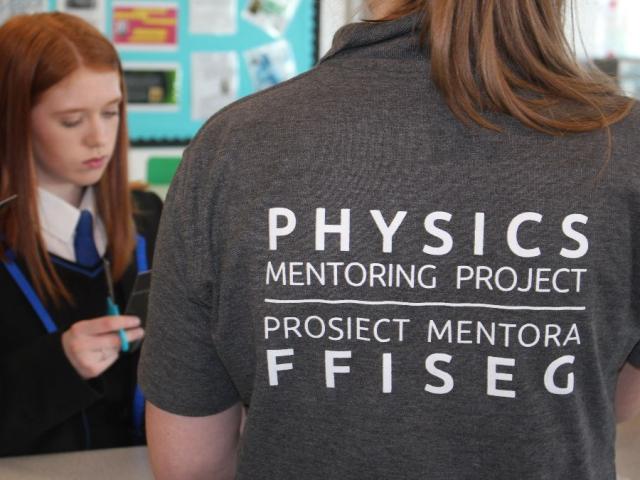 A female school student and another female with her back to the camera wearing a grey t-shirt that says Physics Mentoring Project