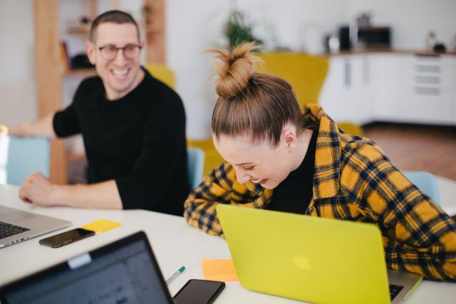 A young man and young woman working at laptops and laughing
