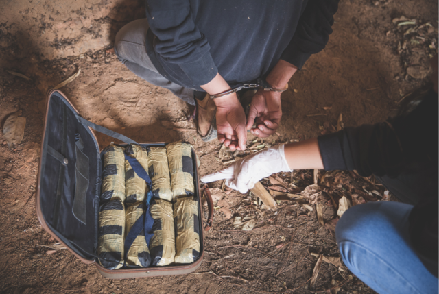 A person in handcuffs kneeling next to a bag of drugs and a person wearing latex gloves pointing at the bag