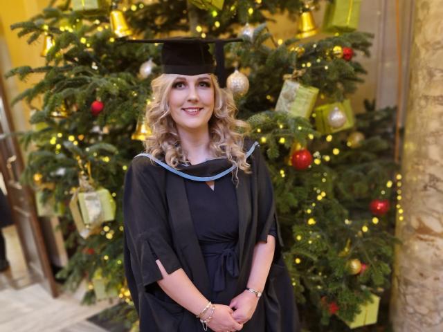 Hayley in graduation cap and gown standing in front of a christmas tree