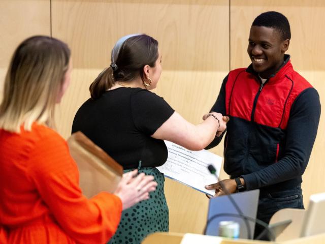 A woman shakes hands with a young man while handing him a certificate. Another woman applauds in the background