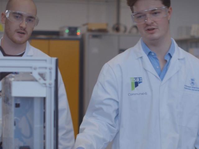 Two men wearing white lab coats, blue gloves and protective goggles, standing in front of a piece of equipment