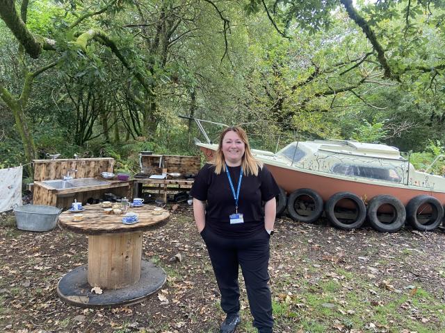 A woman dressed in black standing in a forest setting with wooden benches, wooden table, and a boat behind her